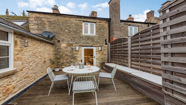 A stone building with a wooden fence, a round table with chairs, and a door leading to an interior space.