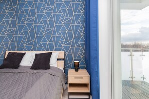 Ein Schlafzimmer mit Wand im geometrischen Muster, einem Bett mit grauer Tagesdecke, zwei Kissen, einem Nachttisch mit Lampe und Blick auf einen Balkon.