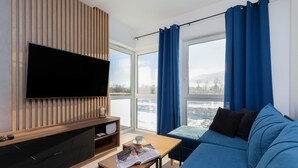 Una sala de estar moderna con un televisor de pantalla plana, un mueble de madera para entretenimiento, un sofá azul, una mesita con una revista y un ventanal con cortinas azules.
