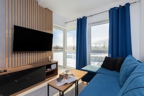 Un salon moderne avec un téléviseur à écran plat, un meuble de rangement en bois, un canapé bleu, une petite table avec un magazine et une grande fenêtre ornée de rideaux bleus.