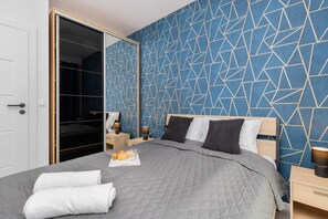 A bedroom with a geometric patterned wall, a bed with grey bedding, two bedside tables with lamps, and a wardrobe with a mirrored door.