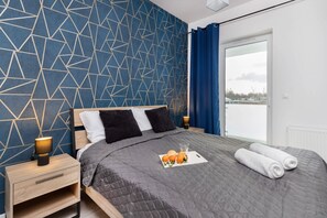 A modern bedroom with a geometric patterned wall, a bed with a gray quilt, two pillows, a bedside table with a lamp, and a tray with oranges and a glass.