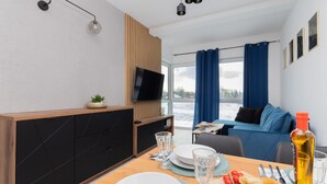 Una habitación de hotel moderna con zona de comedor, televisor, ventana grande con cortinas y vistas al exterior.