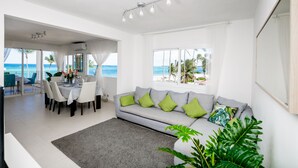 A modern living room with a grey sofa, green cushions, a dining table set for four, and large windows overlooking a beach. - Punta Cana Condo for Rent (Punta Cana)