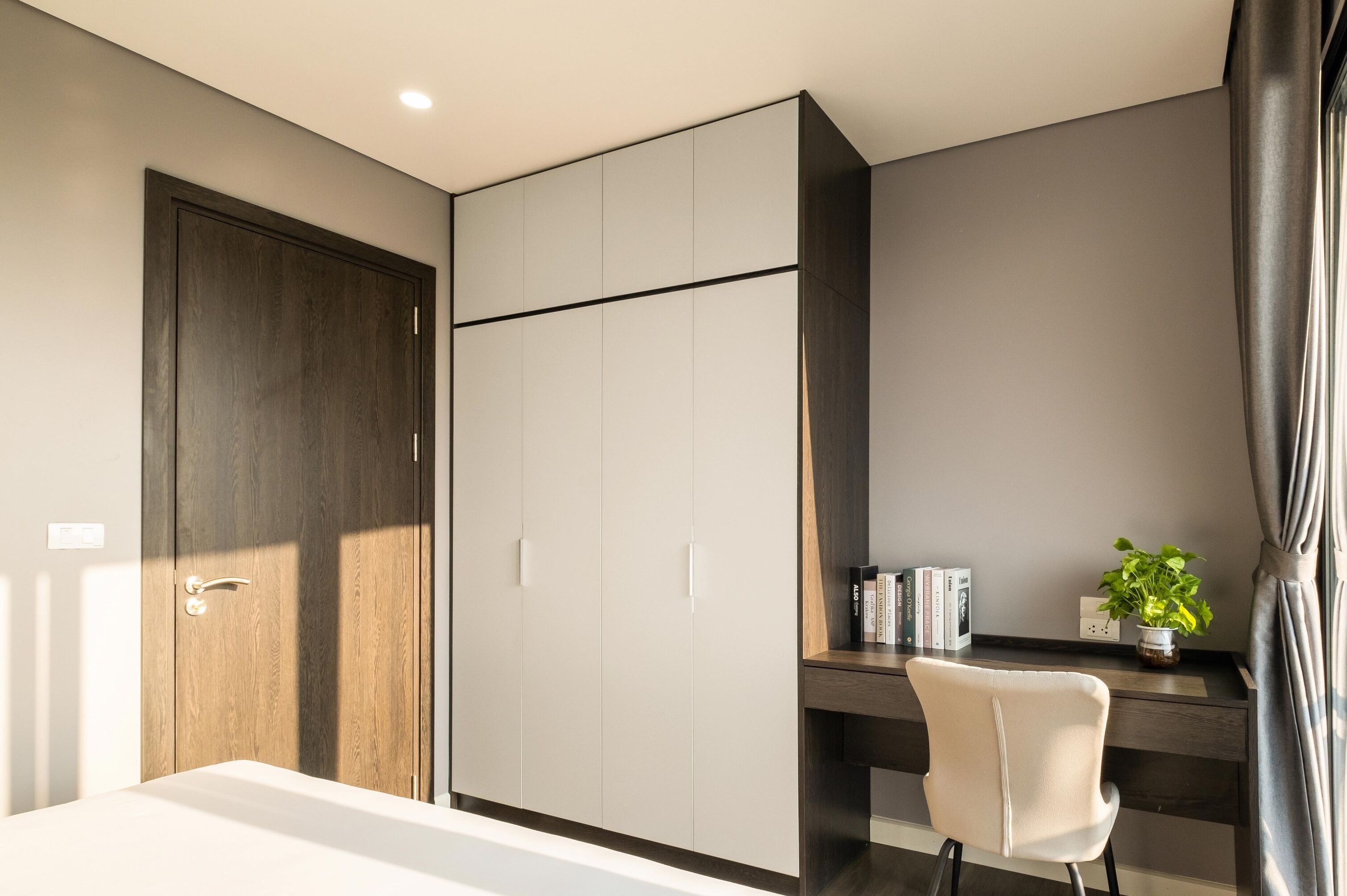 A modern bedroom with a large wardrobe, a desk with a chair, a potted plant, and a bedside table.