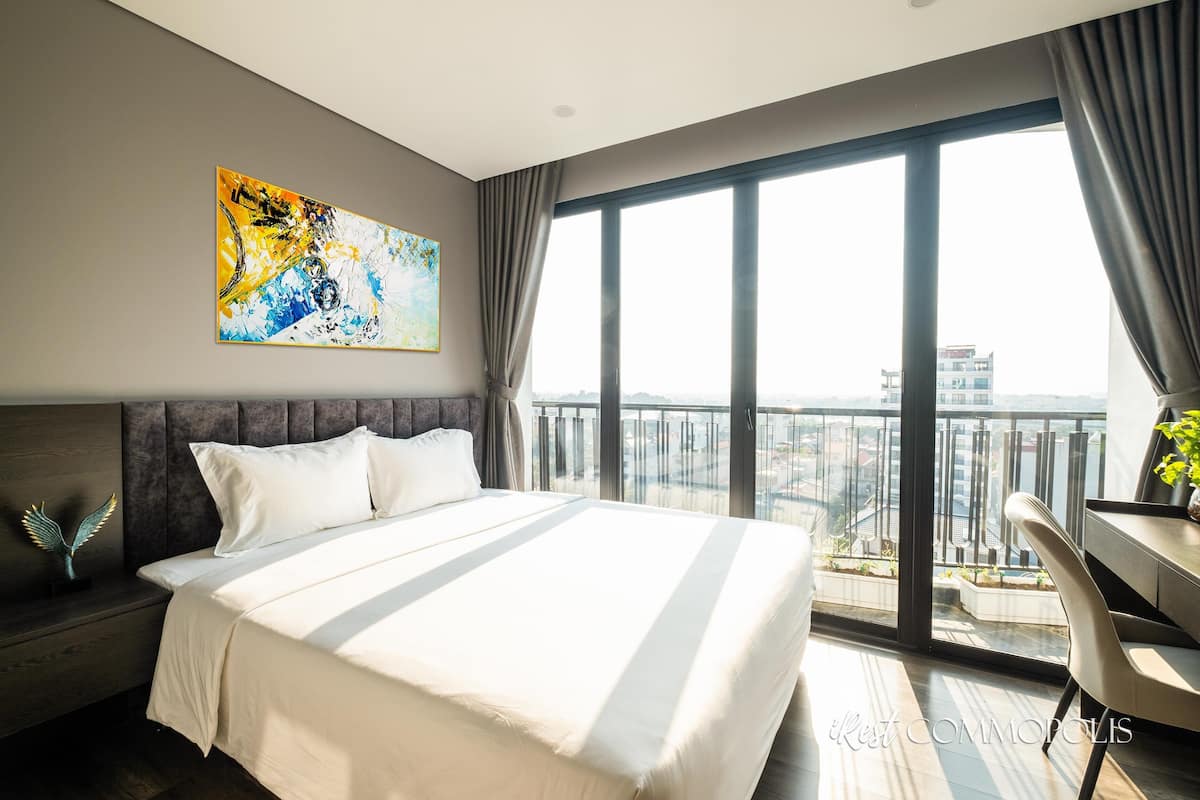 Ett modernt hotellrum med en stor säng, utsikt över staden och en balkong med ett räcke.
