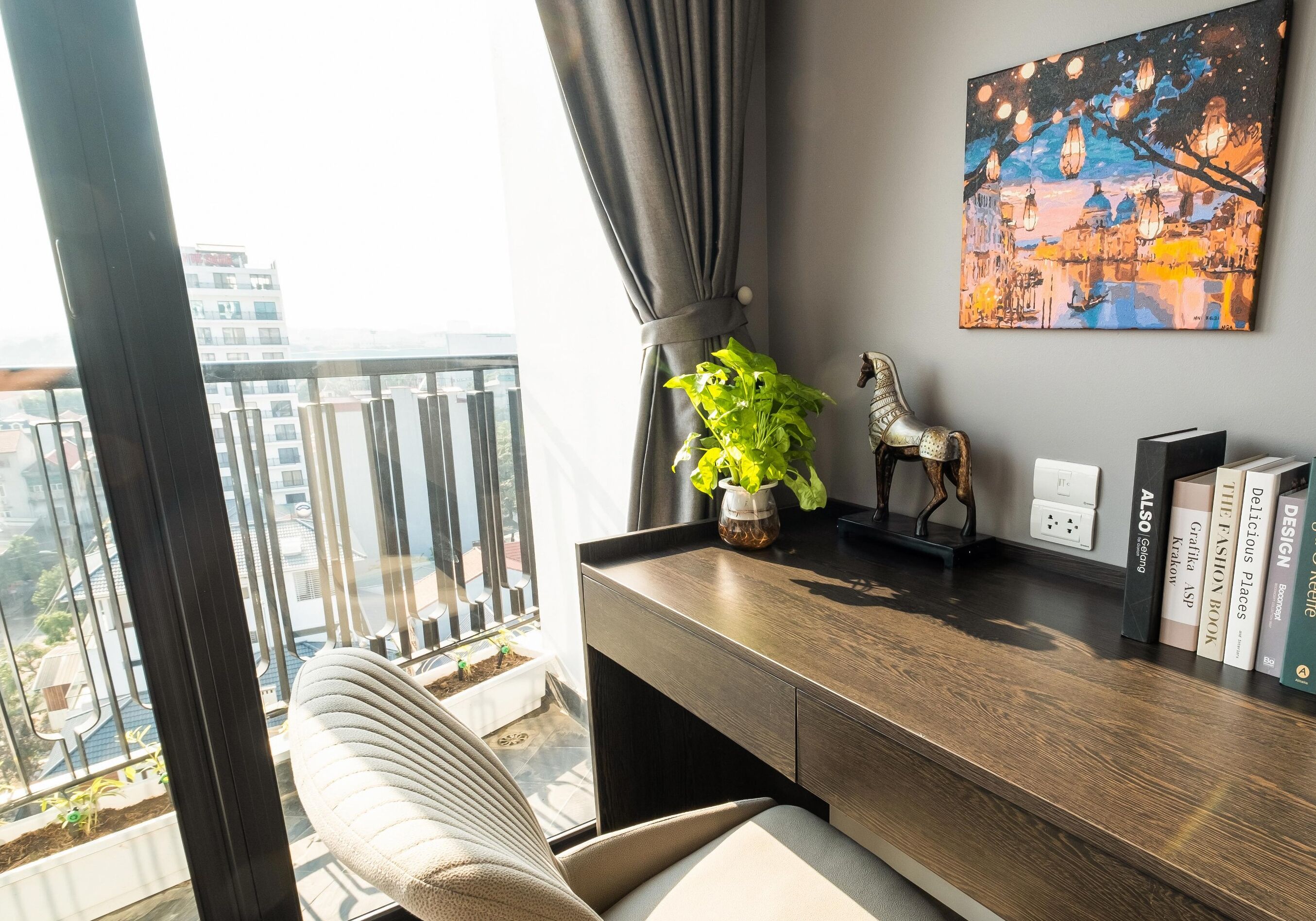 Una sala moderna con vistas a un paisaje urbano a través de amplios ventanales, un sofá cómodo, un escritorio de madera con libros, una planta decorativa y un cuadro colgado en la pared.