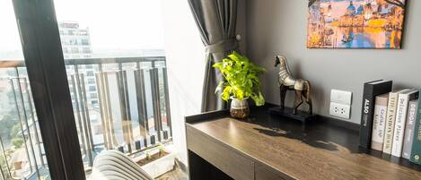 A modern living room with a view of a cityscape through large windows, a comfortable sofa, a wooden desk with books, a decorative plant, and a wall-mounted painting.