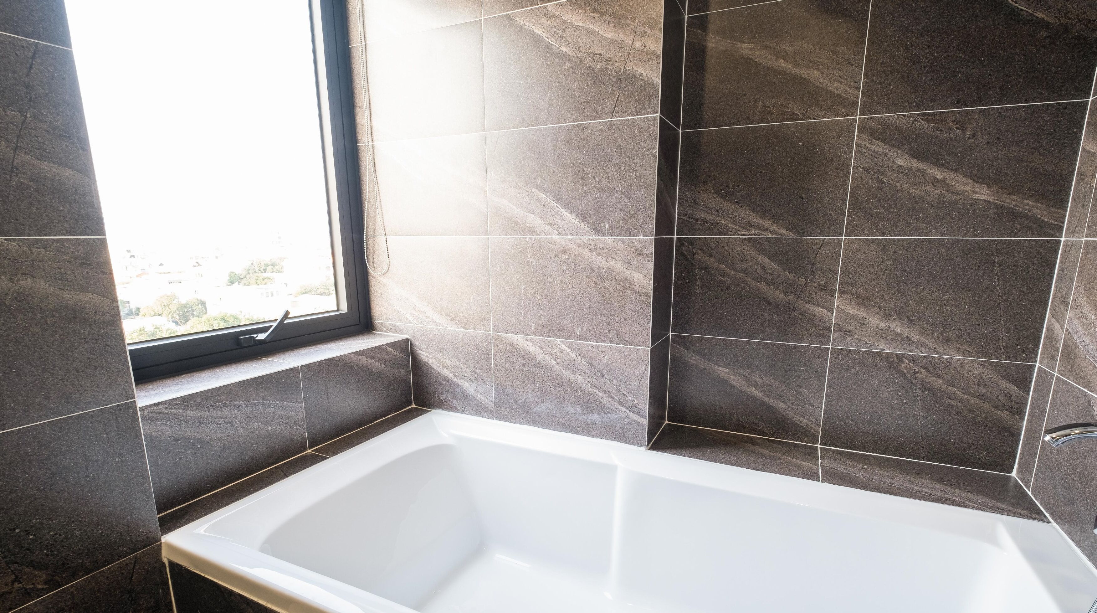 A modern bathroom with a bathtub, a window, and dark tiled walls.