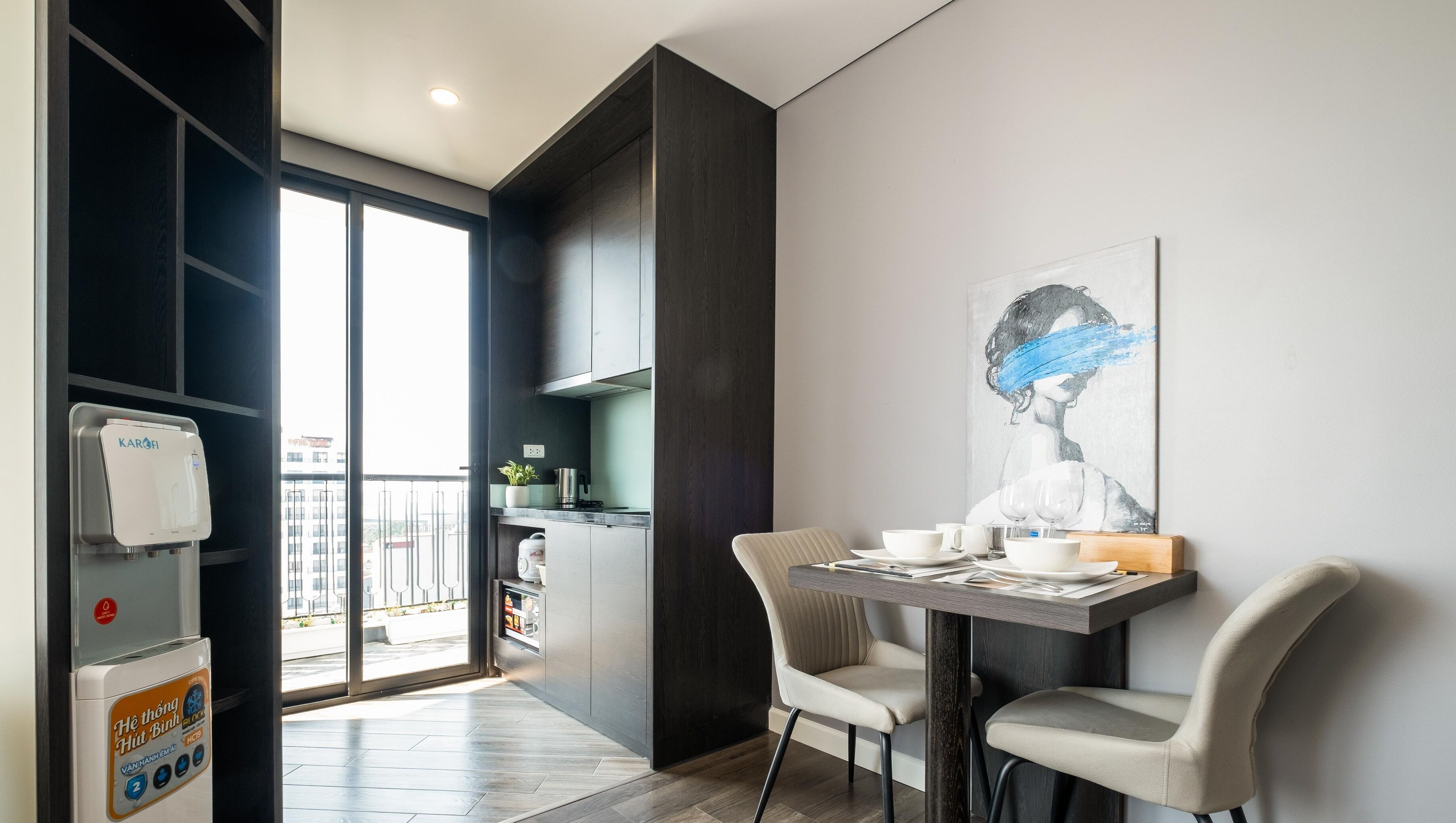 A modern hotel room with a dining area, a water dispenser, a view of the city, and a painting on the wall.