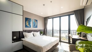 A modern hotel room with a large bed, a desk with a chair, and a view of the city through large windows.