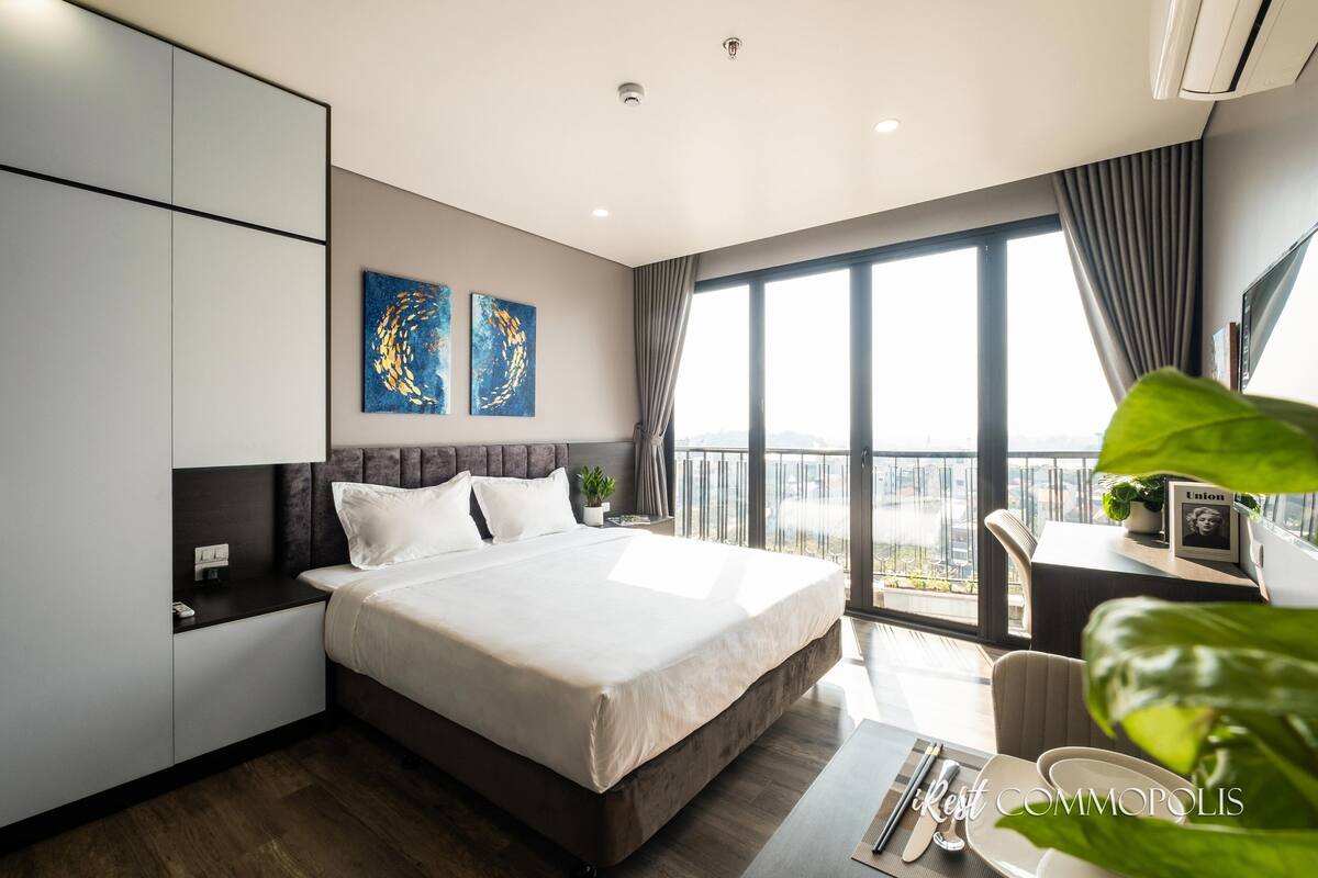 Ett modernt hotellrum med en stor säng, ett skrivbord med en stol och utsikt över staden genom stora fönster.