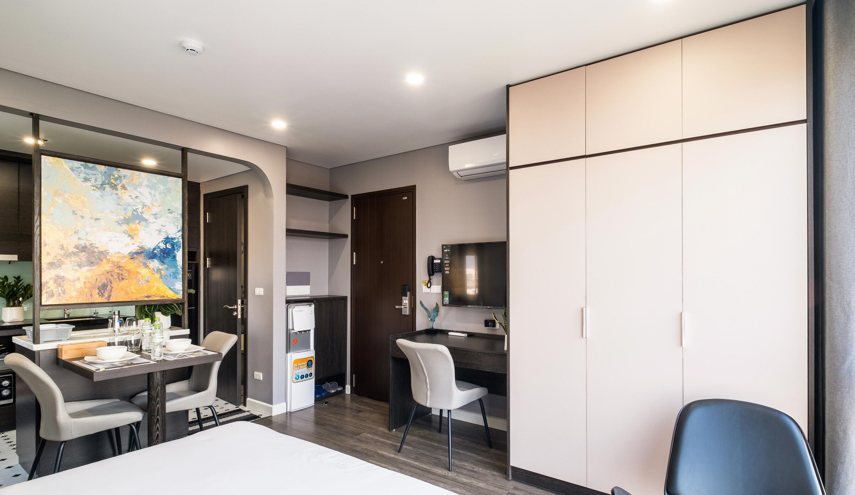 A modern hotel room with a dining area, a large painting, a minibar, and a TV.