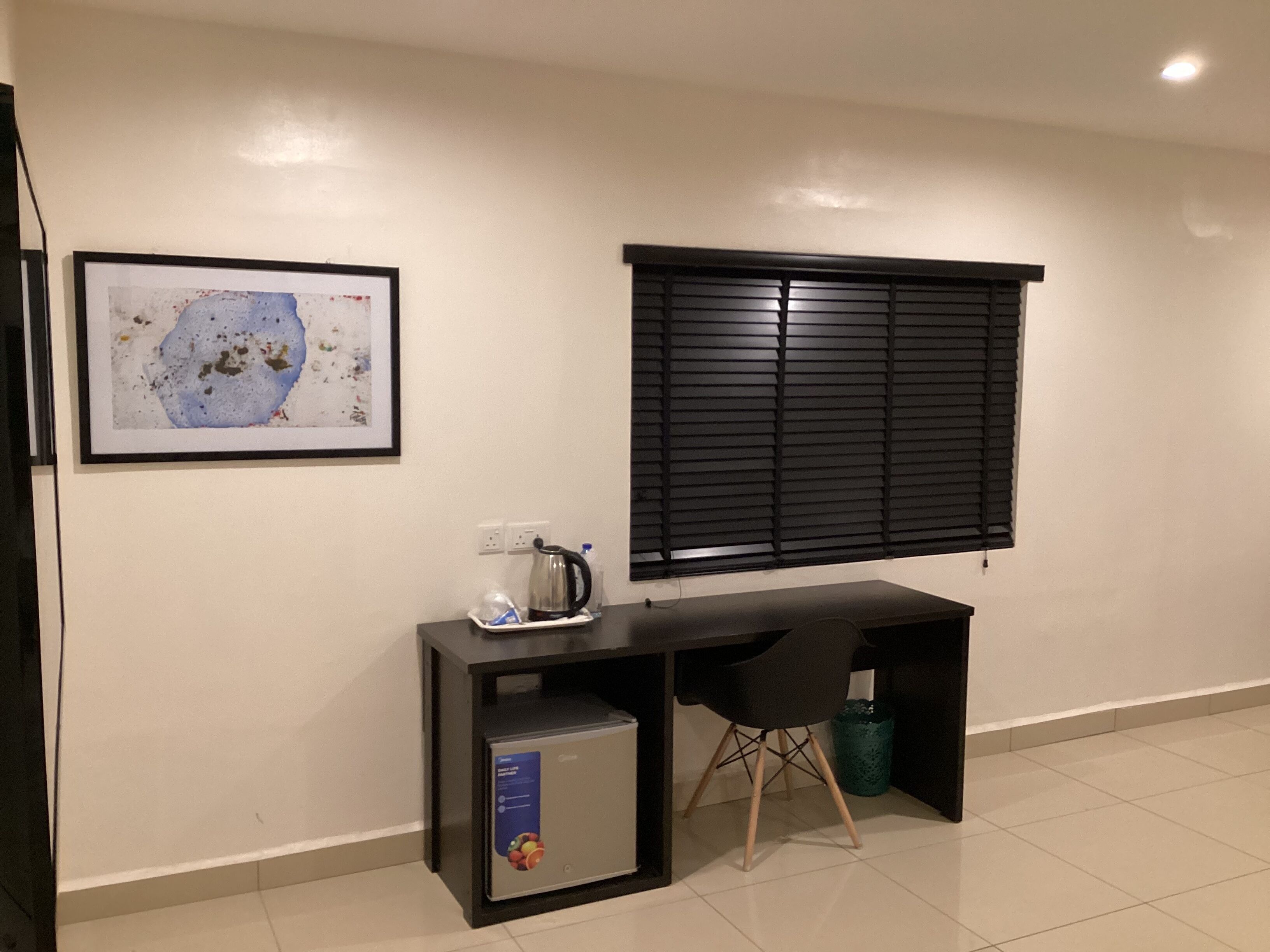 A room with a desk, a window with blinds, a framed artwork, and a small refrigerator.