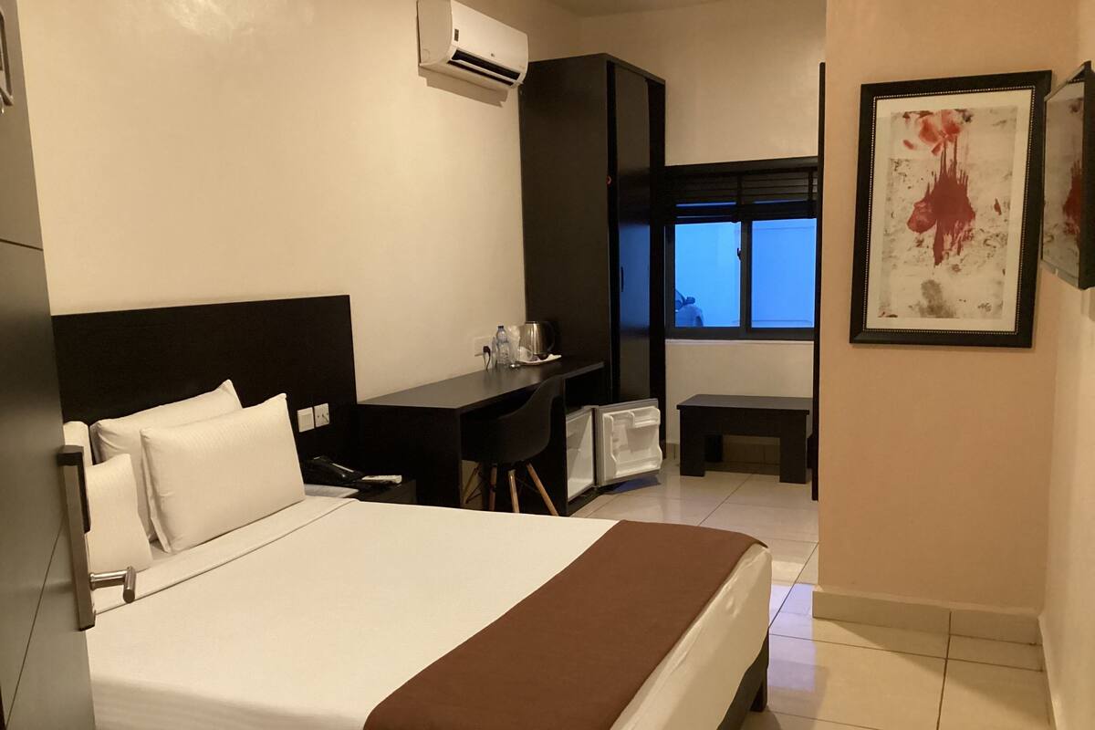 A hotel room with a bed, a desk, a chair, a small table, an air conditioning unit, and a window with a view.