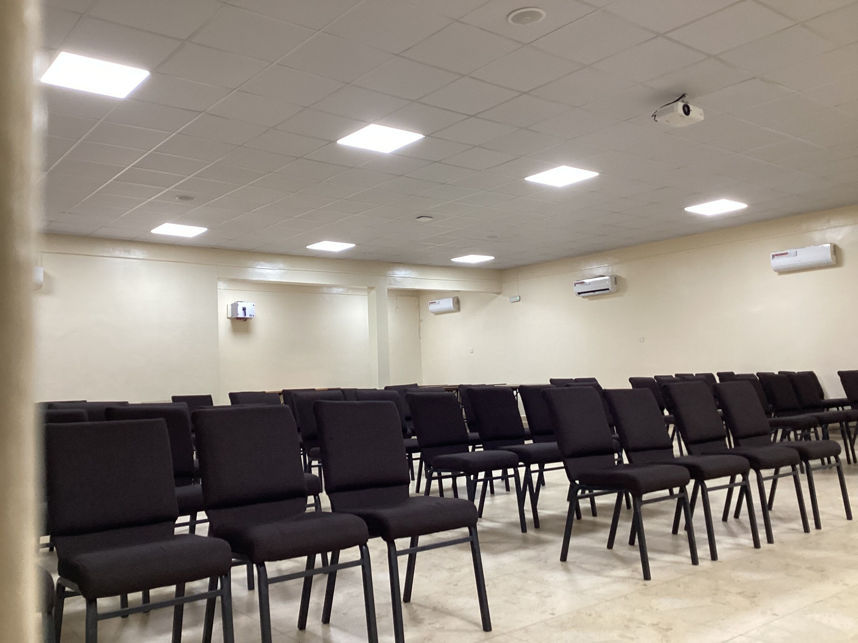 A conference room with rows of black chairs, white ceiling lights, and air conditioning units on the walls.