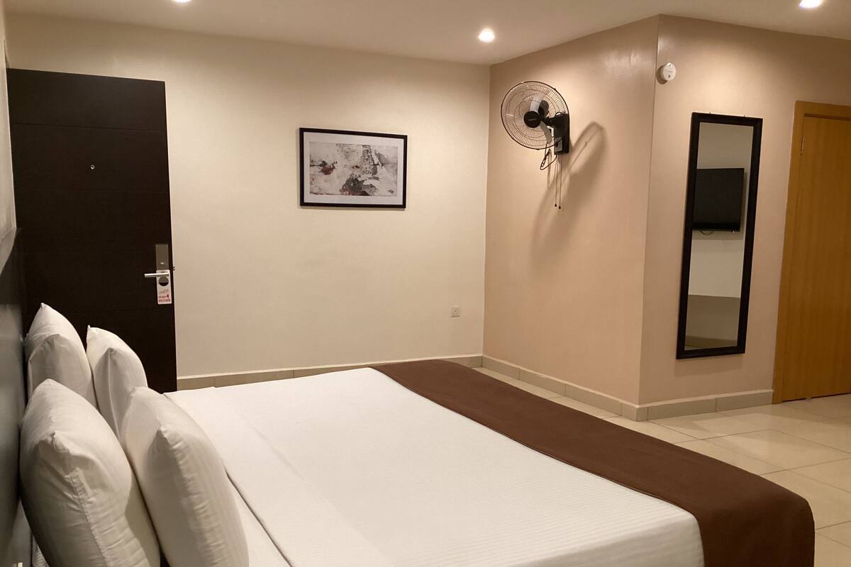A hotel room with a bed, a wall-mounted fan, a framed picture, and a mirror.