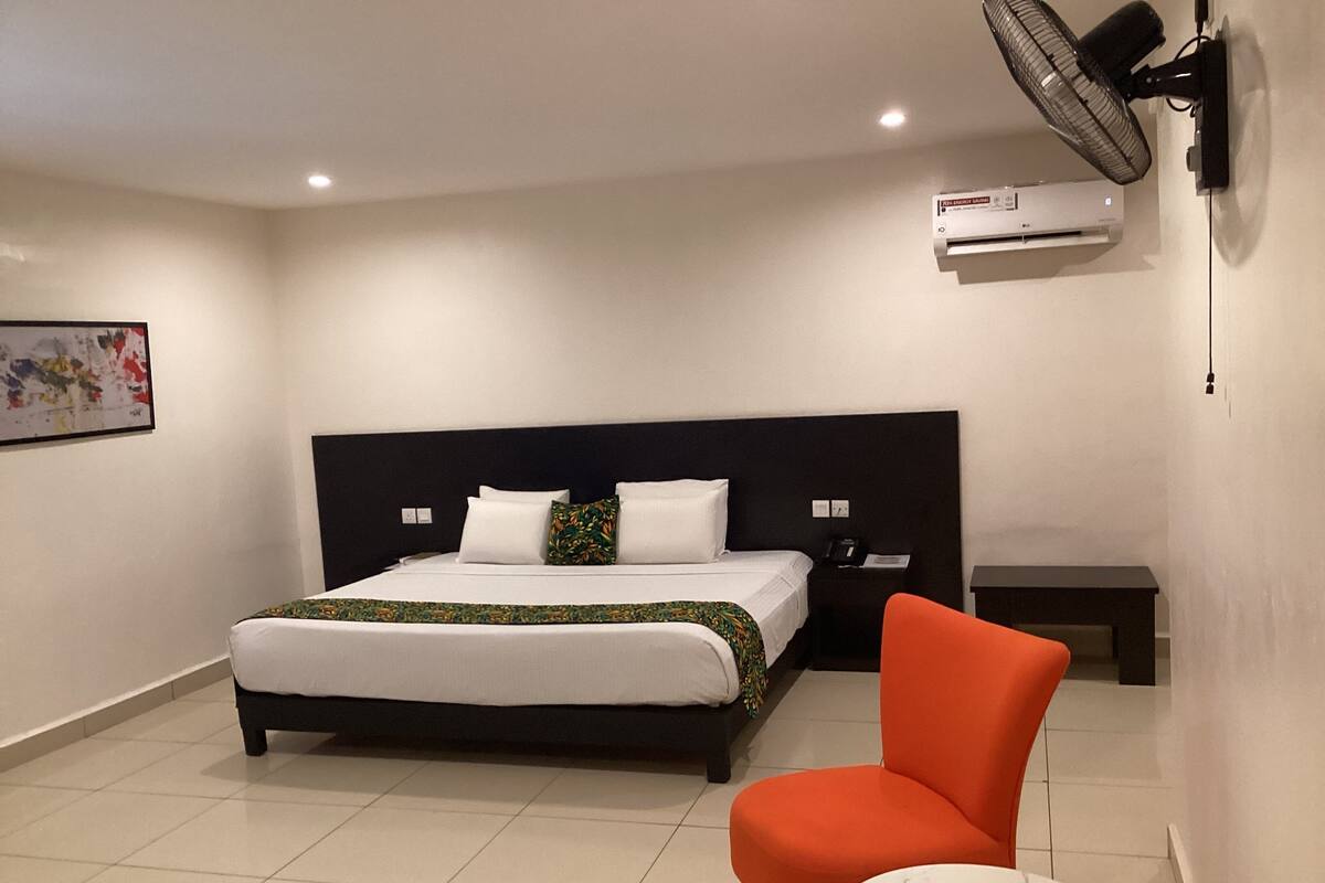 A hotel room with a large bed, a nightstand, a chair, a small table, and a wall-mounted air conditioner.