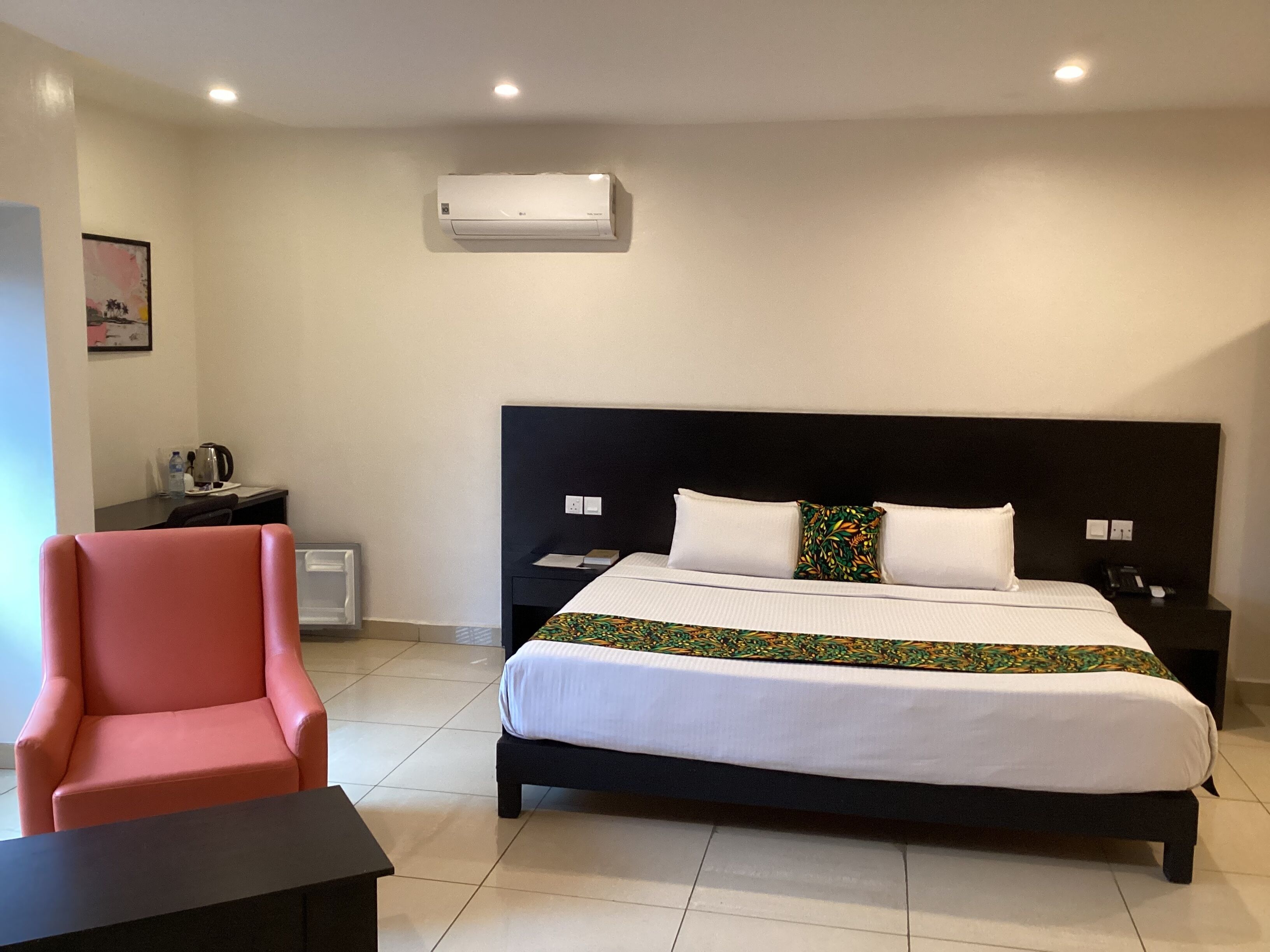 A hotel room with a bed, a chair, a bedside table, a nightstand, and an air conditioning unit.