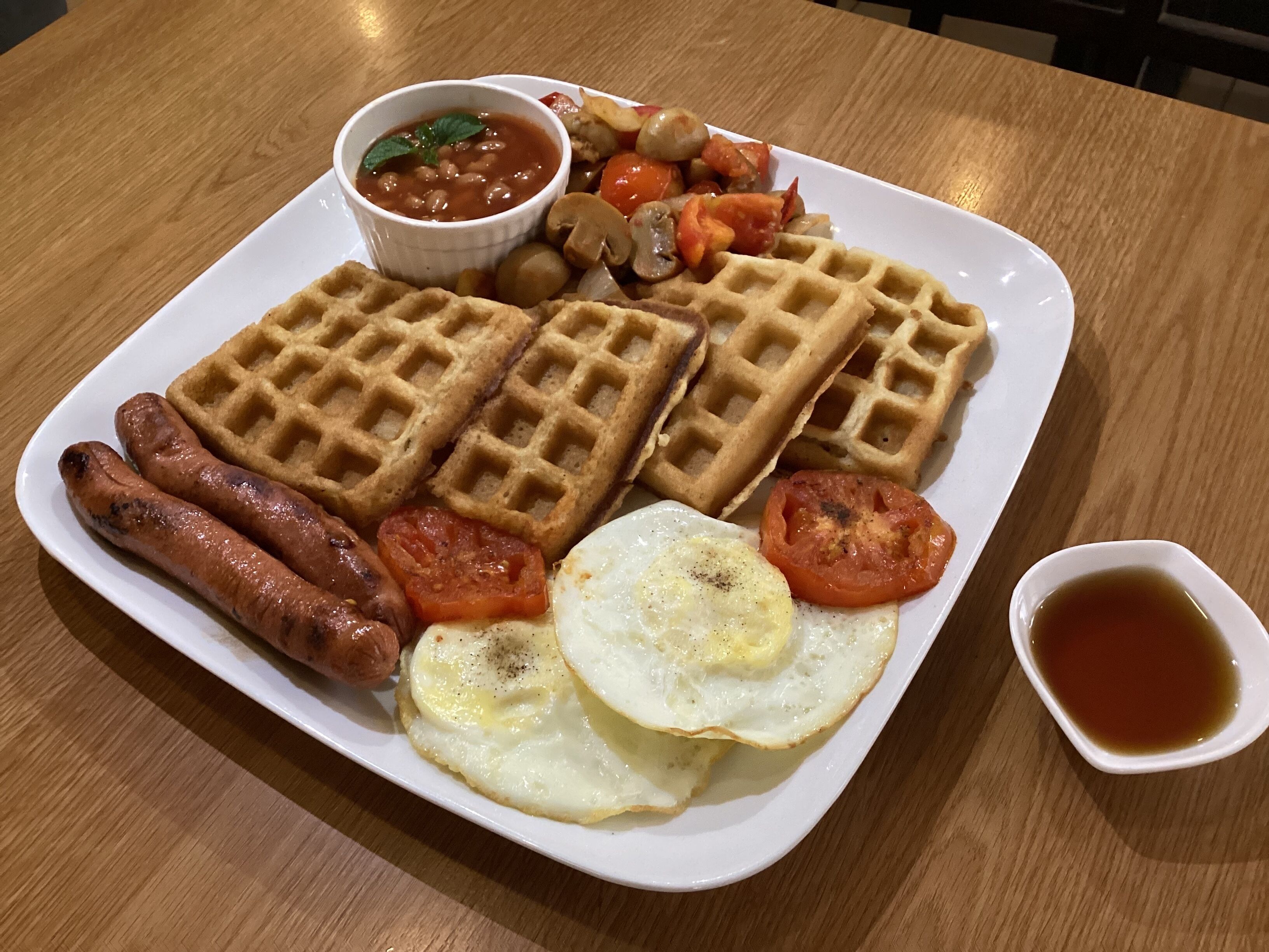 A plate of food including waffles, sausages, eggs, and a small bowl of sauce.