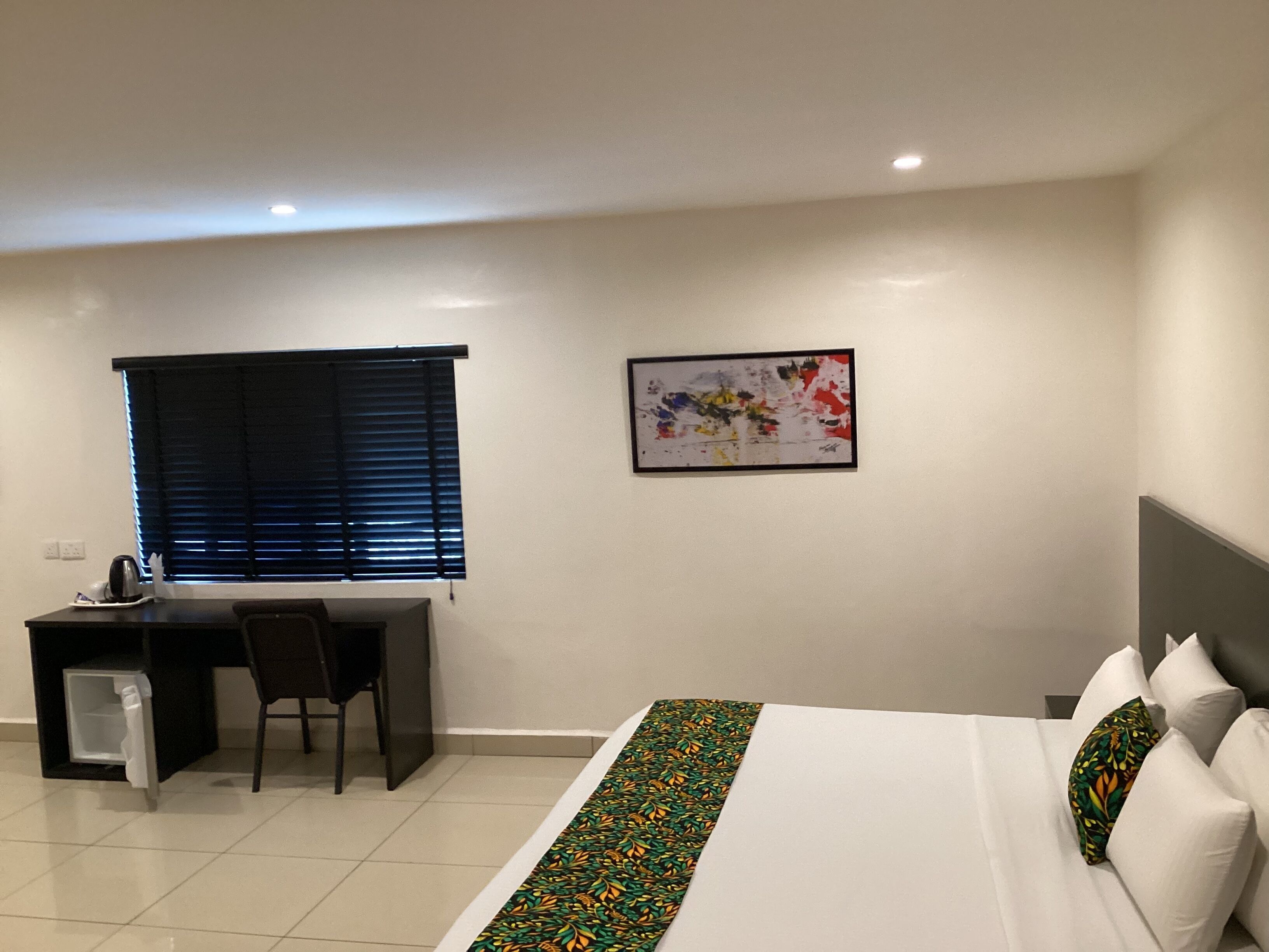 Habitación de hotel con cama, escritorio, silla, ventana con persianas y una obra de arte colgada en la pared.