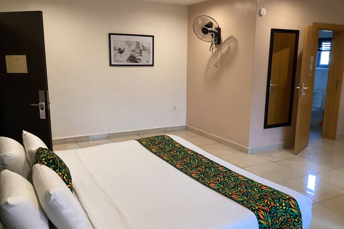A hotel room with a bed, a wall-mounted fan, a door, and a framed picture on the wall.