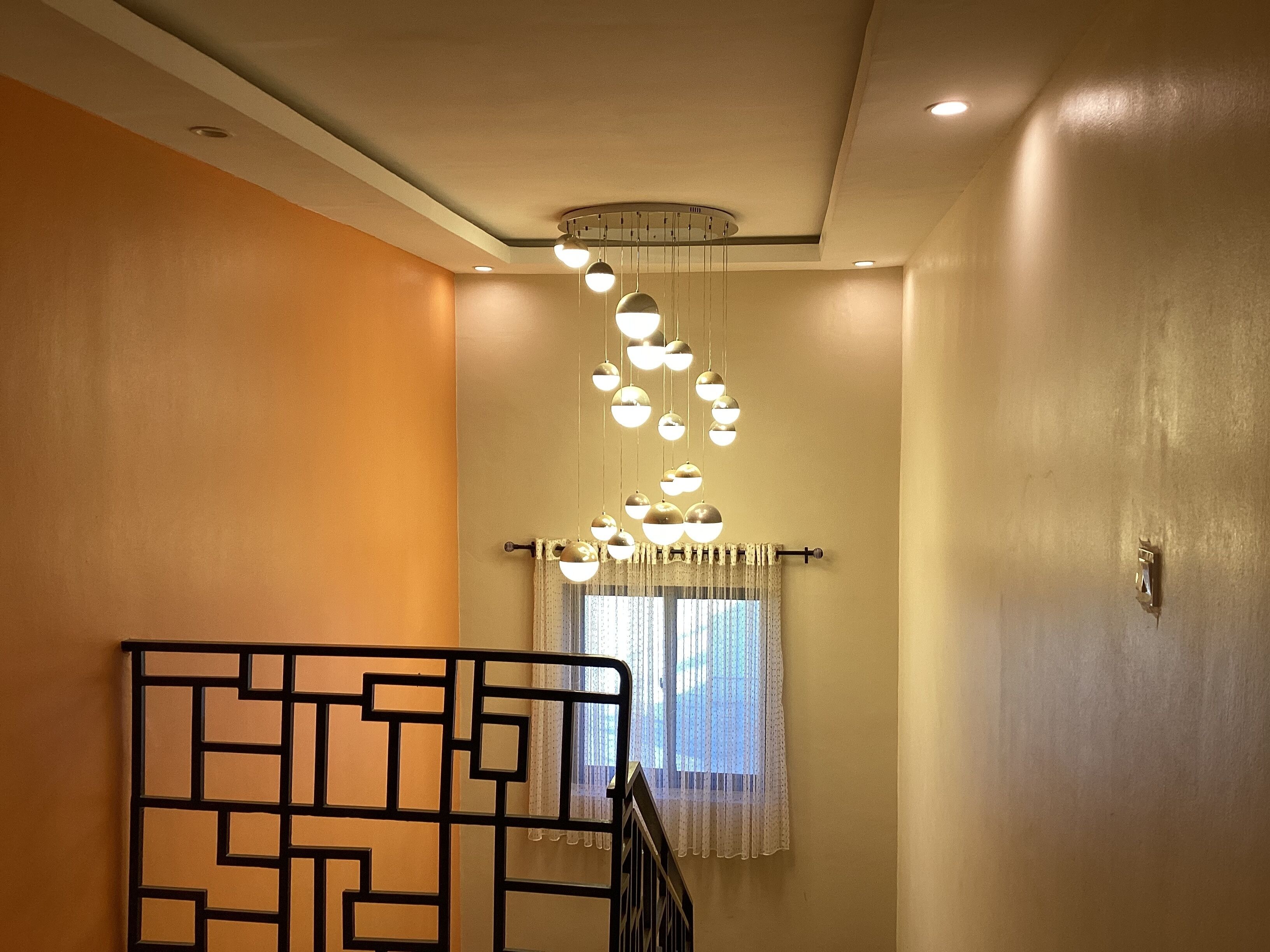 A staircase with a geometric metal railing, a ceiling light fixture, and a window with sheer curtains.