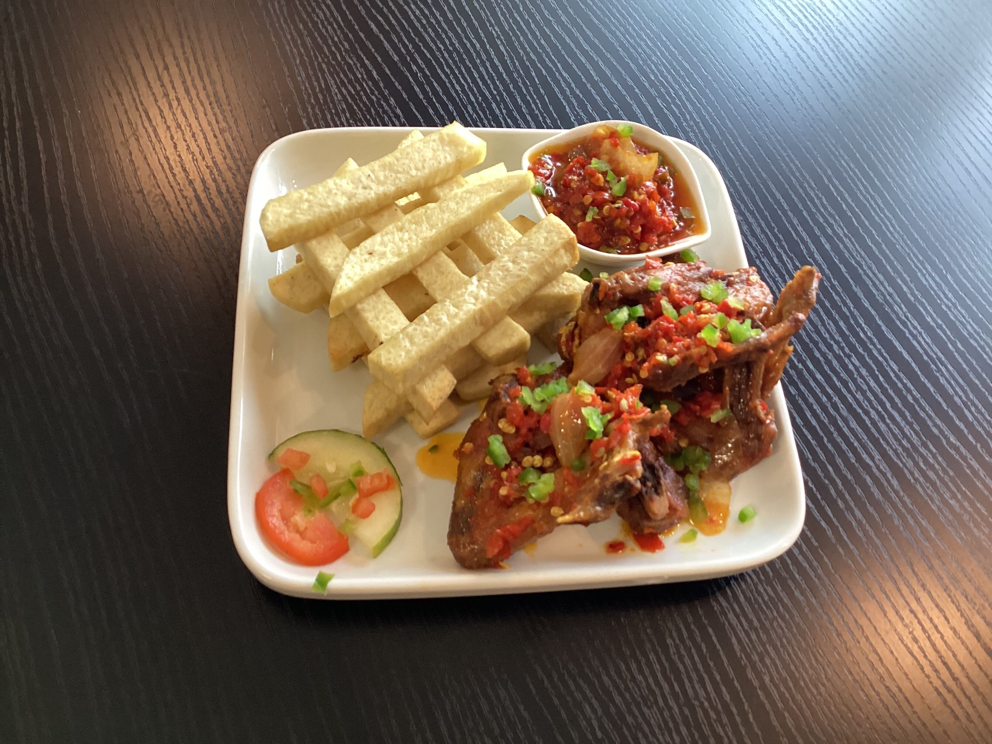 Un plato cuadrado con pollo frito, papas fritas, una rodaja de pepino y un recipiente pequeño con salsa.