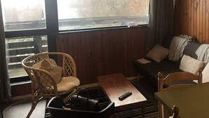 Une pièce chaleureuse avec un décor intérieur en bois, un fauteuil confortable, un canapé, une petite table et une fenêtre donnant sur des arbres et une route.