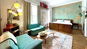 Un salon confortable avec un canapé, une table basse et un lit doté d’une tête de lit en bois. Une carte est accrochée au mur et une plante est posée sur le rebord de la fenêtre.