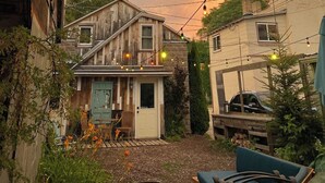 Une maison en bois à l’allure rustique, dotée d’un petit porche, entourée de verdure, avec une voiture visible au loin.
