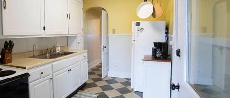 Una cocina con armarios blancos, piso a cuadros, cafetera y un colgador para ollas.