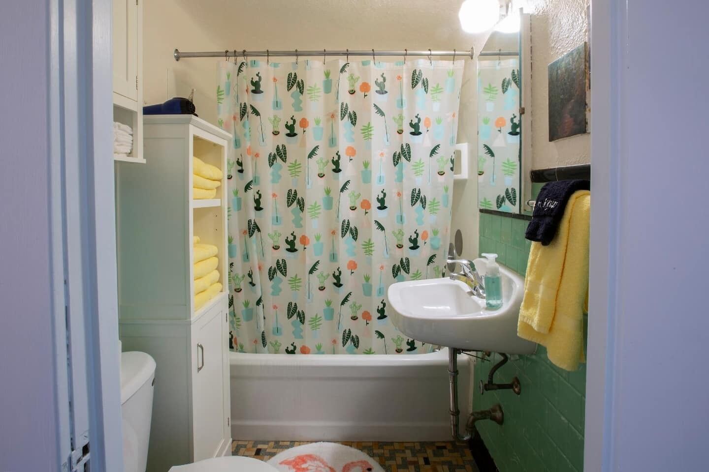 Ein Badezimmer mit Duschvorhang mit Kaktusmuster, einem weißen Waschbecken mit Seifenspender und einer Toilette mit weißem Schrank darüber.