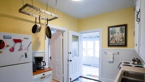 Une cuisine avec des meubles blancs, un carrelage à carreaux, des casseroles suspendues, une machine à café et une porte donnant sur une pièce lumineuse.