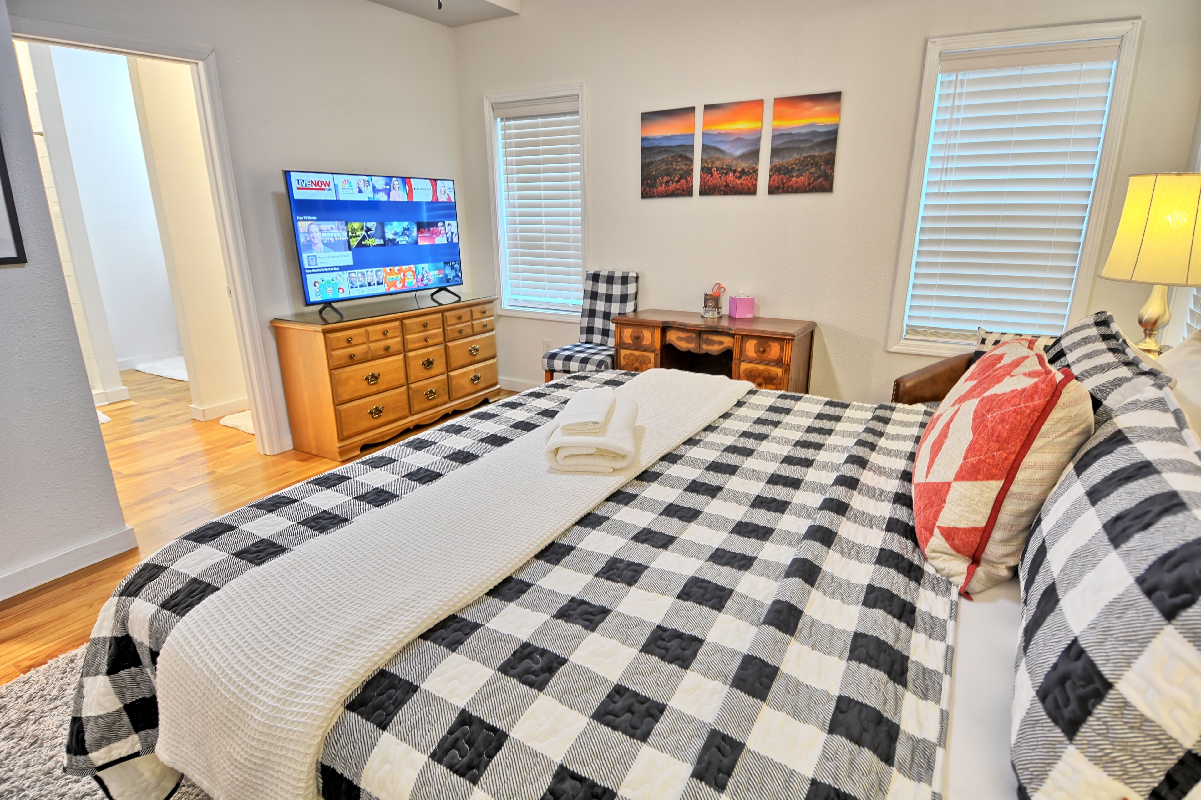 A bedroom with a checkered bedspread, a television on a dresser, a desk with a chair, and a window with blinds.