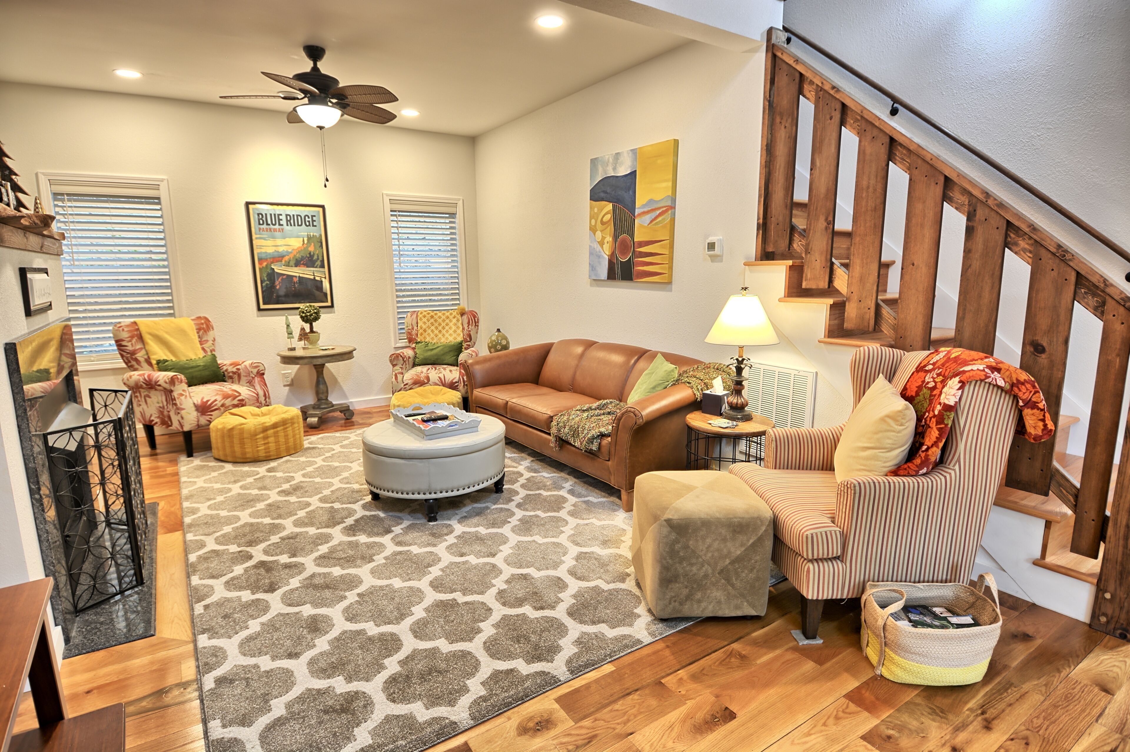 A living room with a brown leather sofa, a round ottoman, a striped armchair, a small table with a lamp, and a staircase with wooden railings.