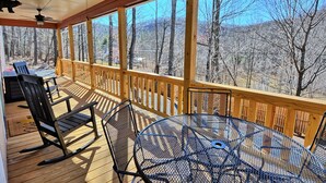 A covered outdoor deck with wooden railings, a table, chairs, and a rocking chair. - Escape to Windy Mountain Dreams in Todd, NC—a cozy chalet for 8 with stunning views! (Todd)