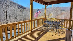 A covered outdoor deck with a table and chairs, an American flag, and a view of a forested landscape. - Escape to Windy Mountain Dreams in Todd, NC—a cozy chalet for 8 with stunning views! (Todd)