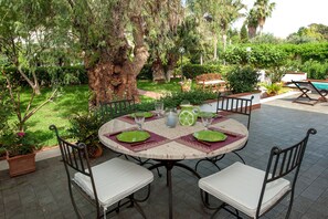 Une table ronde d’extérieur, agrémentée d’assiettes vertes et de coussins blancs, entourée de chaises, est installée dans un jardin où se trouve un grand arbre et diverses plantes.