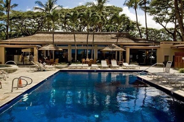 A swimming pool with lounge chairs and umbrellas, surrounded by palm trees and a building with large windows. - Remodeled, Ground floor studio at The Mahana #102 (Lahaina)