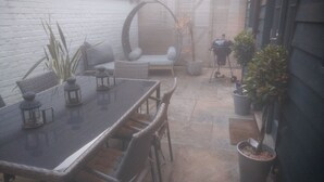 Un espace patio comprenant une table à plateau de verre, des chaises en osier, un coin salon avec des coussins et des plantes en pot.