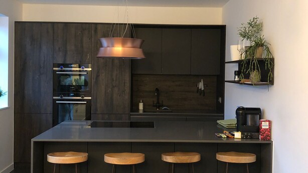Une cuisine moderne avec des meubles de couleur sombre, un îlot central doté de tabourets, des appareils électroménagers intégrés et un luminaire suspendu.
