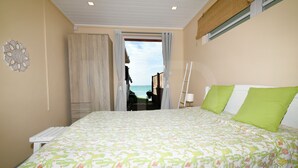 A bedroom with a bed, a window with curtains, a ladder, and a view of the sea. - Beachfront MEERENA, 2 bedroom cottage with Jacuzzi in Calodyne (Calodyne)