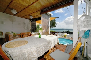 A covered outdoor dining area with a pool, wooden furniture, and a view of the ocean.