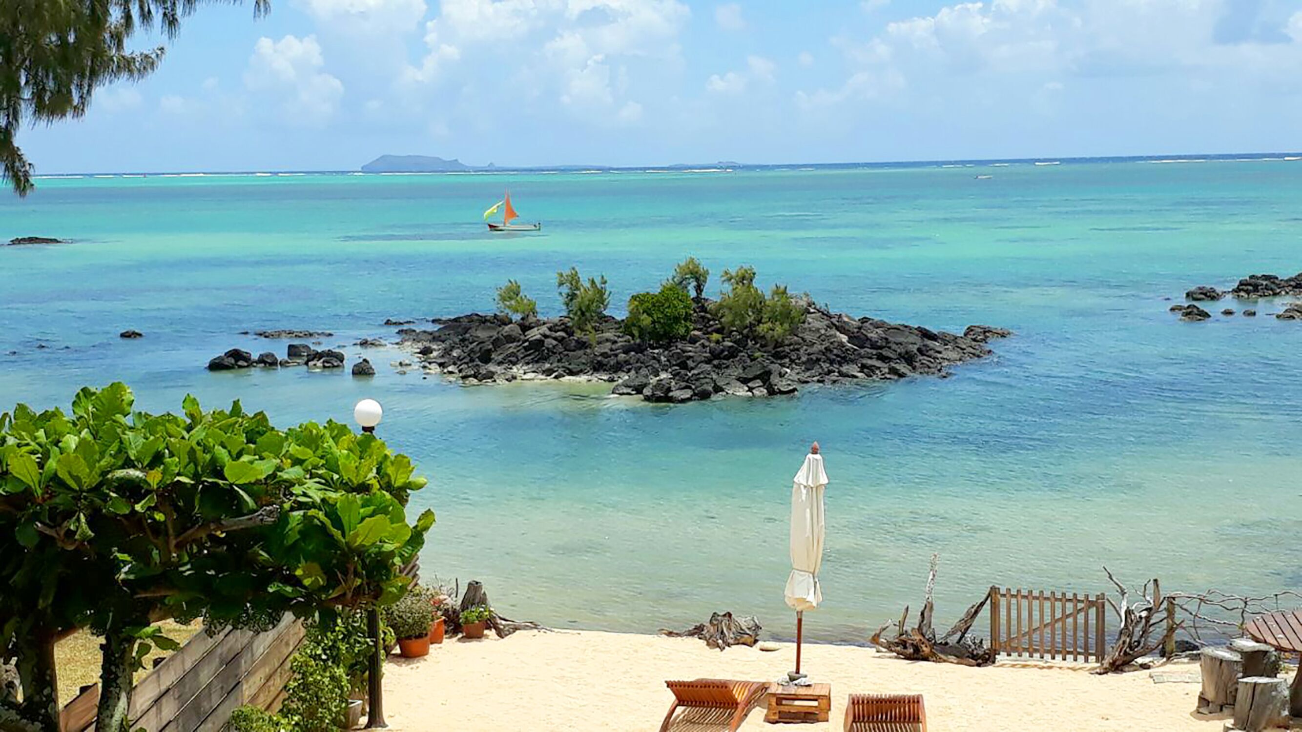 A beach with a rocky outcrop surrounded by clear turquoise water, a sandy area with lounge chairs and an umbrella, and a distant sailboat.