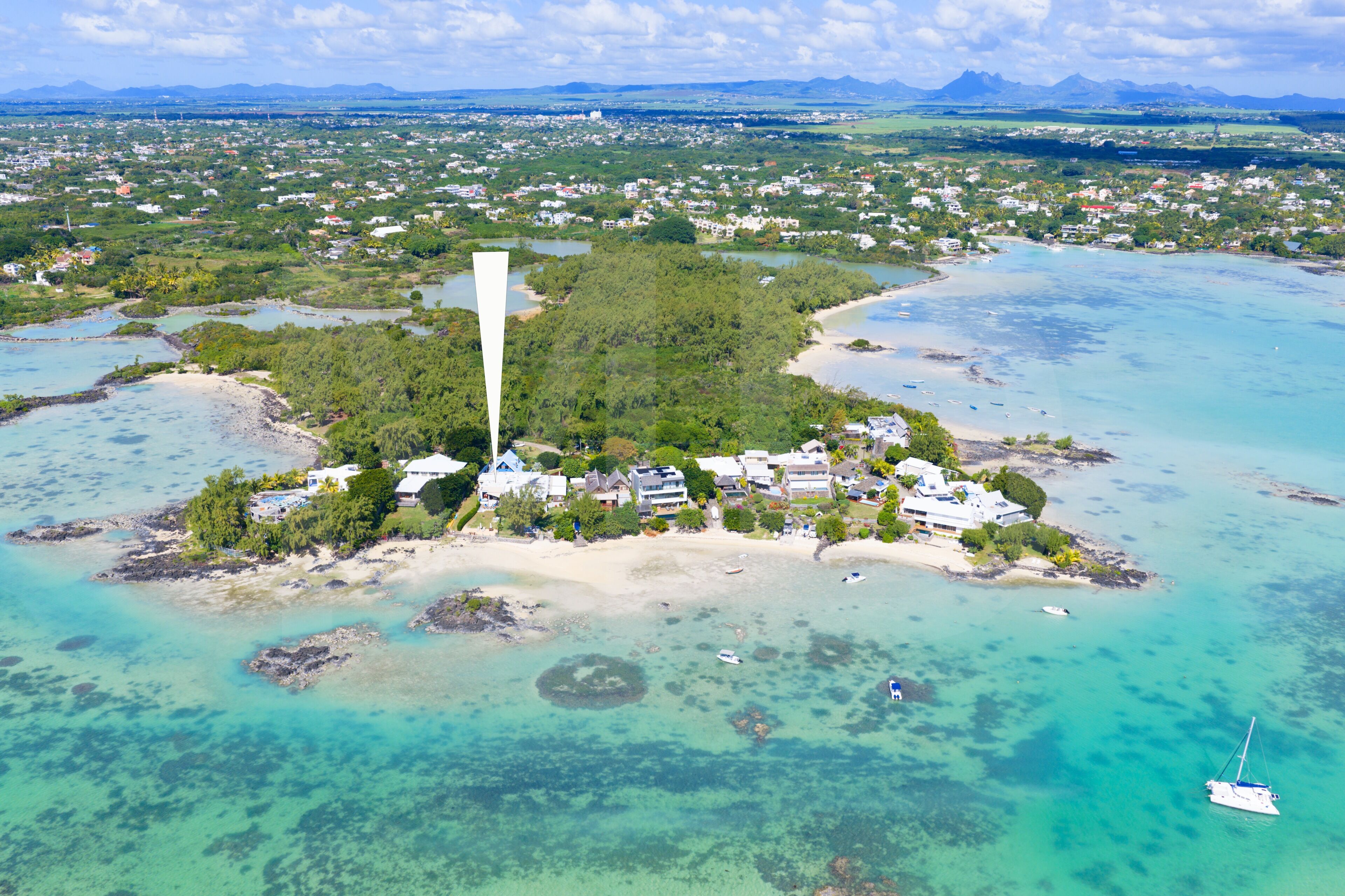 An aerial view of a coastal area with a prominent white tower, a small cluster of buildings, and clear turquoise waters.