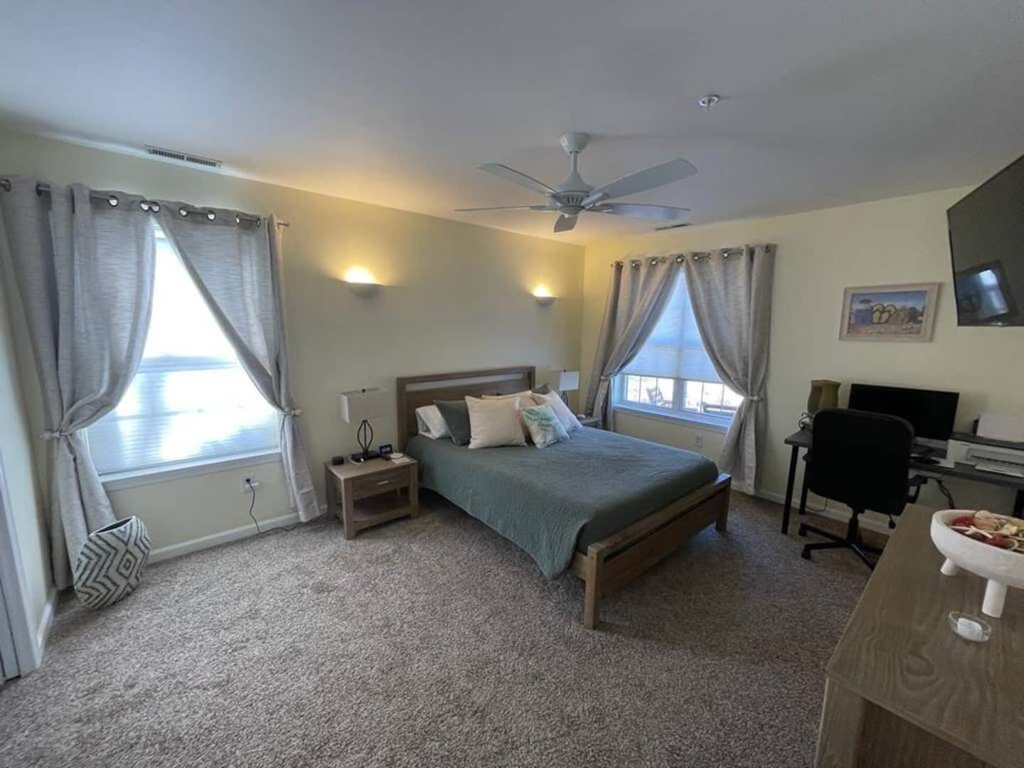 A bedroom with a bed, two bedside tables, a desk with a chair, a ceiling fan, and a window with curtains.