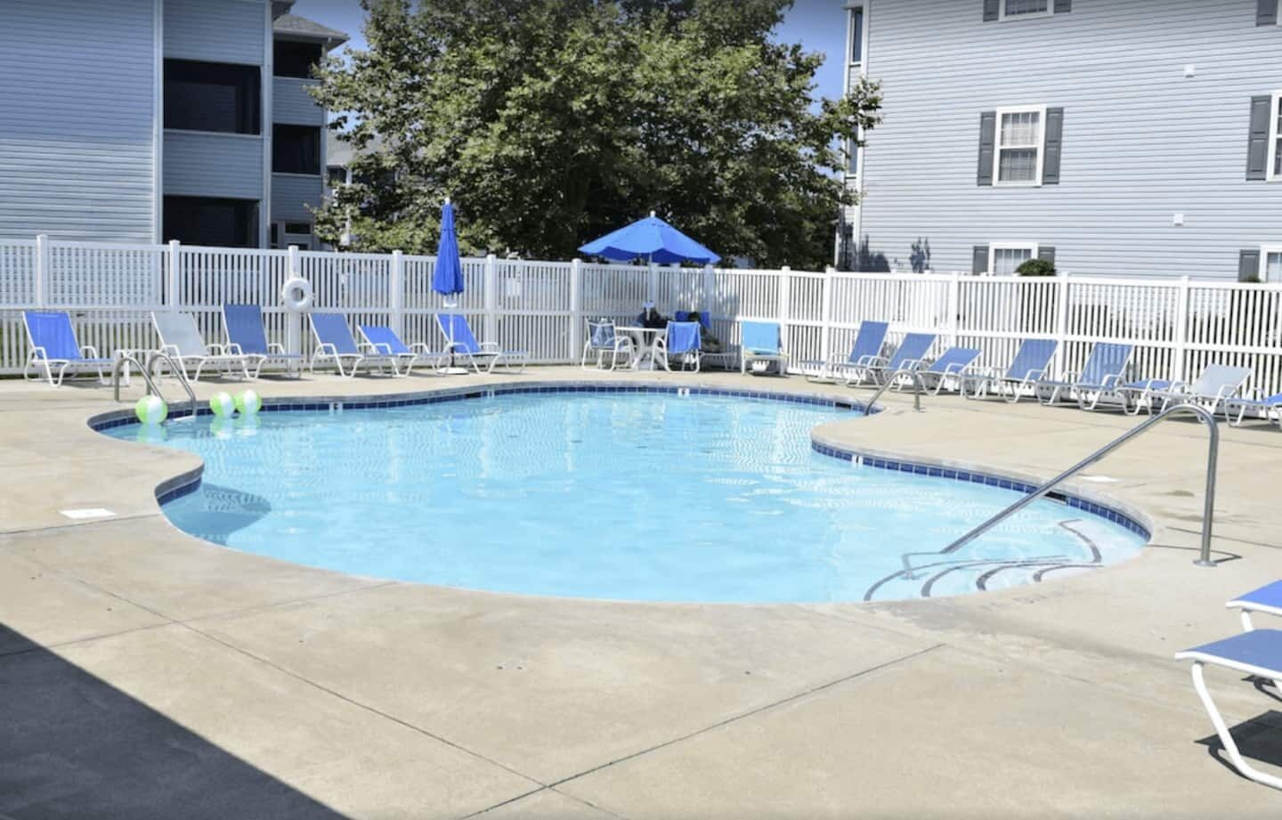 Une piscine rectangulaire entourée d’une clôture blanche et agrémentée de chaises longues bleues.