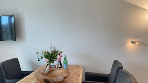 A wooden dining table with a unique bowl centerpiece, flanked by black chairs, in a room with a mounted TV and a wall-mounted lamp. - Strandvilla Wohnung Strand-lodge (Sylt)
