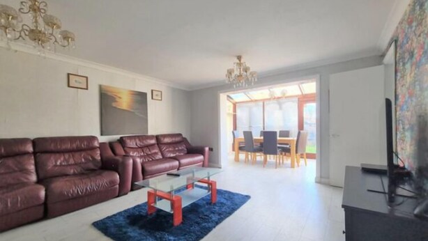 A modern living room with a leather sofa, a glass coffee table, and a blue rug. There is a dining area with a table and chairs visible through an open door.